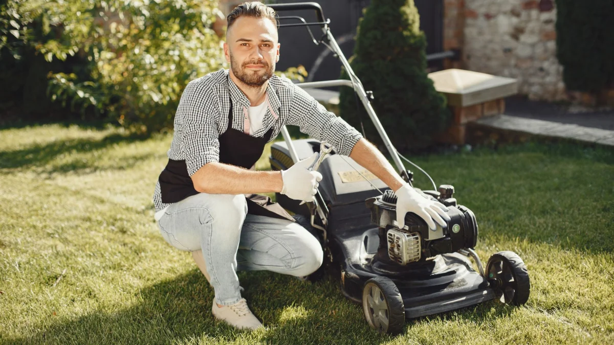 Volwassen man in een schort en handschoenen onderhoudt een grasmaaier op een zonnige dag.