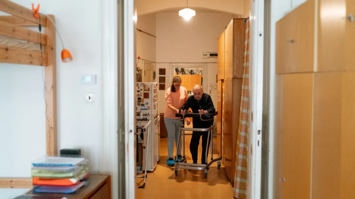 Een verzorger helpt een oudere man met een rollator in een comfortabele thuisomgeving.